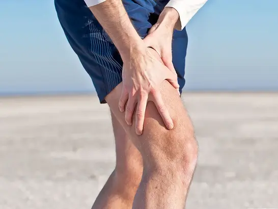 Eine Person steht am Strand und hält mit beiden Händen ihr linkes Knie. Sie trägt ein weißes Langarmshirt und blaue Shorts. Im Hintergrund sind Sand und ein blauer Himmel zu sehen.