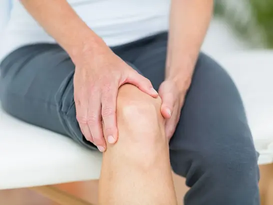 Eine Person sitzt auf einer weißen Fläche, trägt dunkle Hosen und berührt mit beiden Händen ihr Knie. Das Bild suggeriert mögliche Knieschmerzen oder Beschwerden.