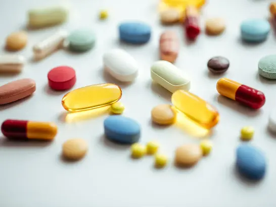Eine Vielzahl bunter Pillen und Kapseln, die auf einer weißen Oberfläche verstreut sind, darunter rote, blaue, gelbe und grüne Tabletten sowie durchscheinende gelbe Gelkapseln.