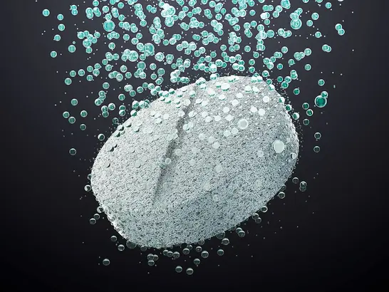 Eine Nahaufnahme einer sich in Wasser auflösenden Tablette, die vor einem dunklen Hintergrund von zahlreichen kleinen Bläschen umgeben ist.