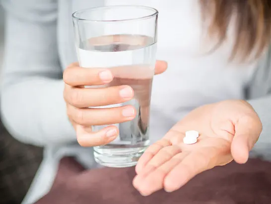 Eine Person, die in einer Hand ein Glas Wasser und in der anderen zwei weiße Pillen hält und ein graues Langarmhemd trägt.
